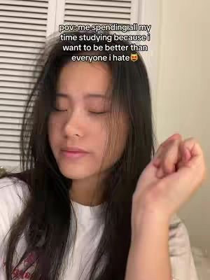 A young woman with long dark hair and a white shirt is positioned in front of a cream-colored wall with vertical lines. The photo is a close-up shot of her face, with her eyes closed. She is holding up her right hand to the side as if she is talking.