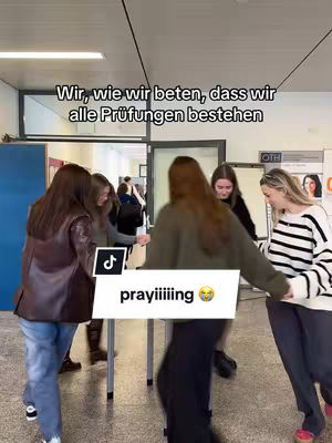 A group of young women are standing around a small table in a hallway, seemingly praying or huddling for good luck. The image has a natural, candid feel. Text overlays on the image.