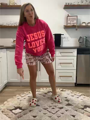 A woman wearing a pink sweatshirt and shorts stands inside a kitchen setting, pointing downwards with her right arm. The sweatshirt has the words "JESUS LOVES YOU" printed on it in orange, and she is wearing slippers. The background includes white cabinets, a stainless steel appliance, and some decorative items on shelves.