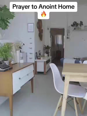 An interior shot of a well-lit home. The camera is positioned to capture a corner of a room, with a wooden desk and white drawer unit on the left. A white chair and the corner of a light-colored wooden table are visible on the right. In the background, a doorway leads to another room. The wall behind the desk is white, complemented by plants. The overall impression is clean, bright, and inviting.