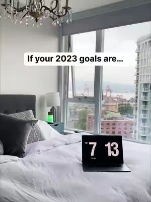 A bedroom scene with a large window overlooking a cityscape. A bed with white bedding and gray pillows is in the foreground. A laptop displaying the time '7:13' rests on the bed. A text overlay reads 'If your 2023 goals are...'. A chandelier hangs from the ceiling.