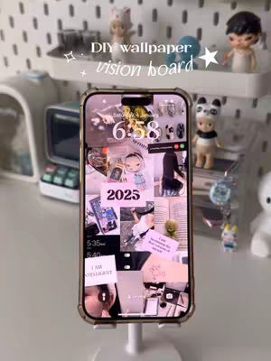 The cover image features a smartphone held upright, showcasing a customized vision board. The phone is positioned on a white stand. A display shelf is behind the phone, and a table is in the foreground. The phone's background is a collage of images. The text overlay 'DIY wallpaper vision board' with stars on top, is displayed on the top of the image. The setting looks aesthetic, clean, and organized.