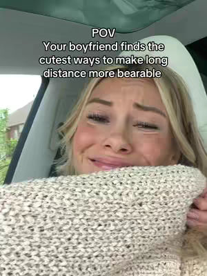 A young woman with blonde hair is seated in a car, visible from the shoulders up. She is wearing a cream-colored sweater. Her face is streaked with tears. A text overlay at the top reads, "POV Your boyfriend finds the cutest ways to make long distance more bearable."