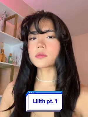 A woman with long dark hair is centered in a close-up shot. She's looking up slightly to her left with a nearly neutral expression. A text box overlay reading "Lilith pt. 1" is in front of her. The background is softly lit, with shelves visible to the left.