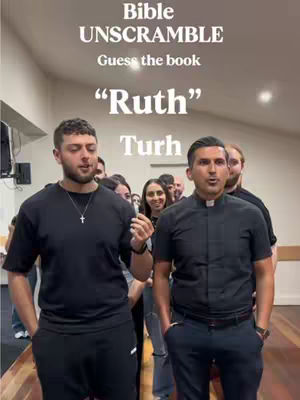 Two men and group of people standing, playing a word game. The man on the left is wearing a black T-shirt and a necklace, the man on the right is in a priest's collar.