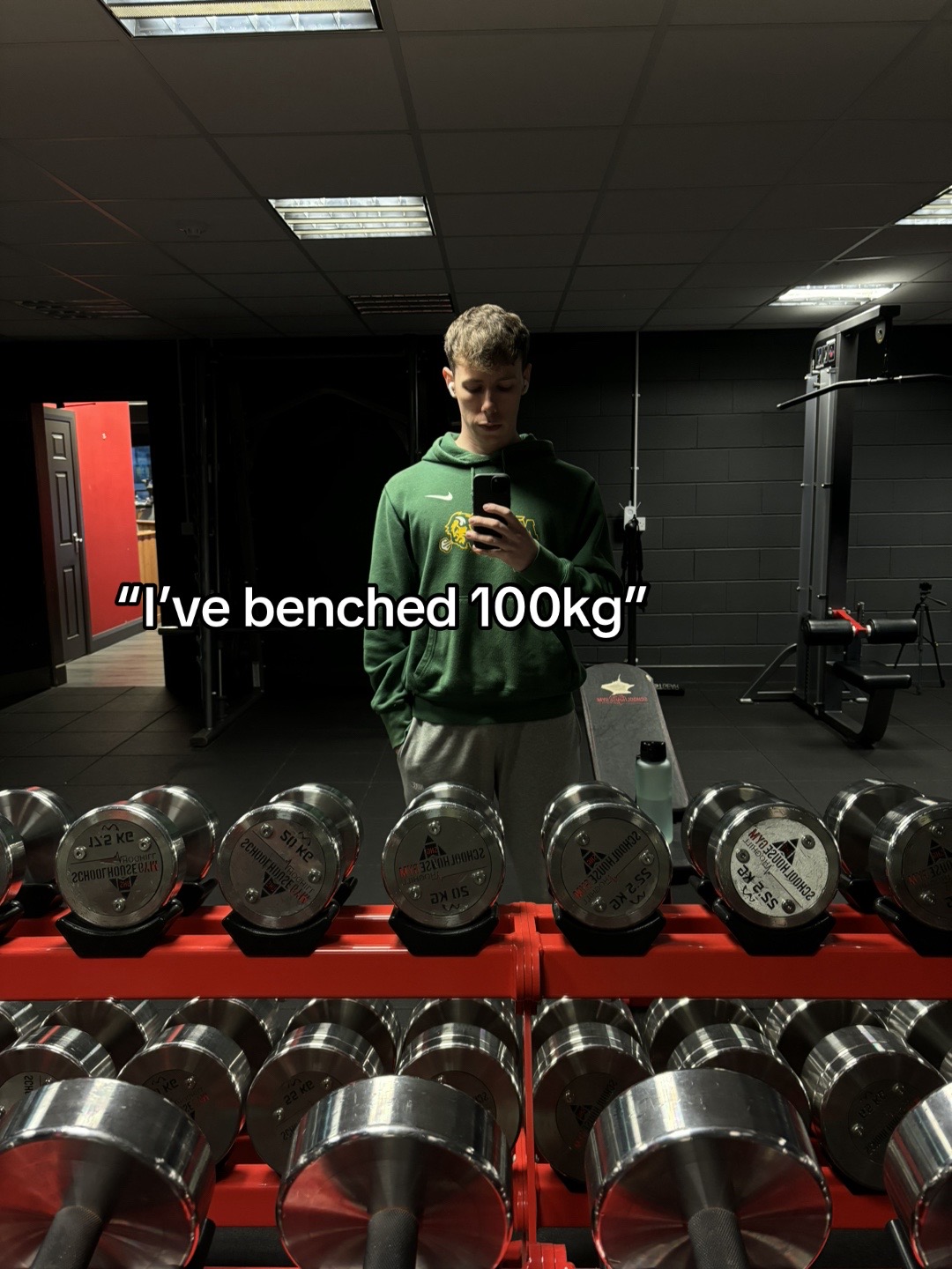A man stands in a gym, centered, holding a phone up to take a mirror selfie. He is wearing a green hooded sweatshirt, grey sweatpants. Dumbbells are in front of the man. The background features gym equipment, including a weight bench and a dark-colored wall. Text overlayed on the image reads, "I've benched 100kg".