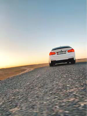 A white BMW car is captured from the rear, positioned on an asphalt road that slopes toward the bottom of the frame. The car is slightly angled, revealing its rear lights and license plate, with a background of a gradient sky transitioning from a golden sunset to a clear blue. The composition is dynamic due to the road's incline.