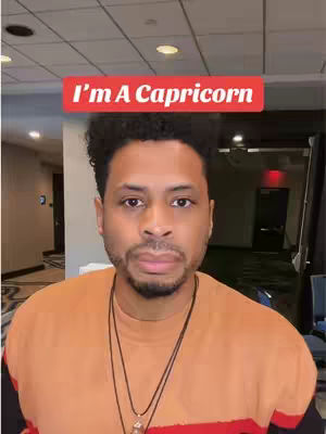 A man with short, dark curly hair and a trimmed beard is the main focus. He wears a two-tone orange and black shirt and a necklace. Above his head, a red banner reads 'I'm A Capricorn'. The background suggests a conference room with a corridor, doors, and a ceiling with recessed lighting.