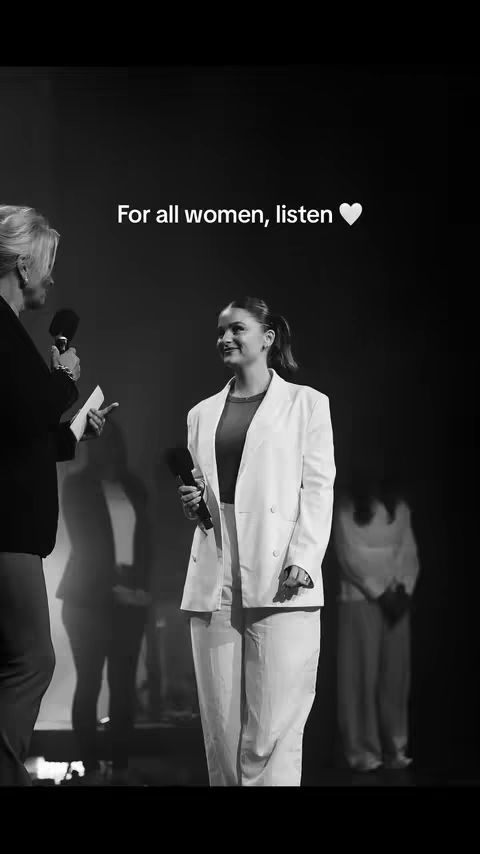 A black and white image shows two women on a stage. The main focus is on a woman in a white blazer, pants, and a dark undershirt, holding a microphone. The woman on the left is partially visible, speaking into a microphone. The background is dark, and there are other figures blurred in the distance, suggesting a live event.