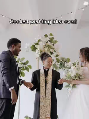 A wedding ceremony is taking place inside a well-lit hall. The central focus is on the officiant, a Black person in a black and gold robe, holding the hands of the bride (right) and the groom (left). The bride is wearing a white wedding dress and holding a bouquet, while the groom is in a suit. Background decorations include large flower arrangements and string lights.