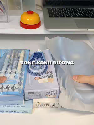 The cover image showcases an organized desk setting with various stationery items. A white shelf holds a silver laptop with a black keyboard, and a yellow bell with a red base sits on the shelf. Below, several blue-themed stationery products are arranged, including pens and packaging, alongside a white box with a blue and white eraser. The composition is well-lit, suggesting a focus on study or desk organization.