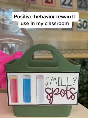 The cover image is a vertical video frame featuring a green container with a handle, holding a sign that reads "Smelly Spots." The sign includes images of tubes of lip gloss in red, blue, and white. The background shows a blurred classroom setting with what appears to be a pink fuzzy item in a glass vase and numbers on the wall.