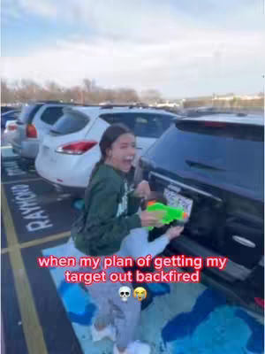 A young woman stands outdoors in front of a black SUV in a parking lot, holding a green water gun and laughing with her mouth open. Text overlaid in the image reads 'when my plan of getting my target out backfired'.