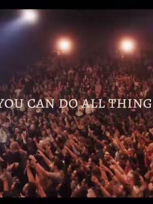 The image displays a large gathering of people, likely at a concert or religious event, with arms raised towards what seems to be a stage. The scene is dimly lit, with spotlights creating a dramatic effect, and text overlayed on the crowd.