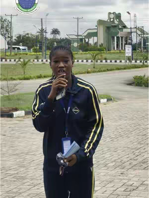 A woman stands in a university setting, holding a phone and papers. The background shows university buildings and landscaping. She's dressed in a dark blue track suit with yellow stripes.