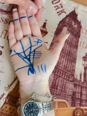 A close-up shot shows a woman's hand, palm up, with blue ink markings resembling lines of palmistry. A silver watch and bracelet adorn the wrist. In the background, a piece of fabric is visible, imprinted with travel-themed images (Big Ben, old car, text labels). Another person’s hand is visible holding the subject’s fingers.