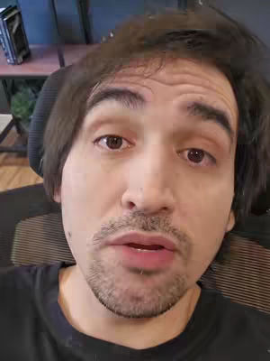 A man with brown hair and a bit of facial hair is looking into the camera, as if talking. The background is blurred but appears to be an office or home setting.