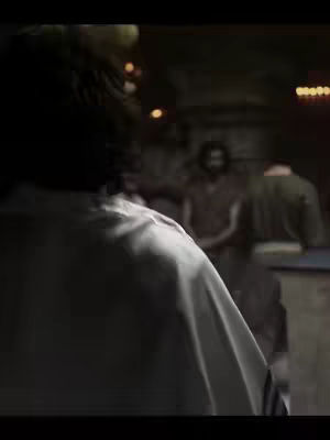 The cover image is a dimly lit interior shot. The primary focus is on the back of a person wearing white clothing, the figure is turned away from the camera. In the background are two other people, with softer focus, one facing the camera and another in profile. Dim lighting and earthy tones create a solemn atmosphere.