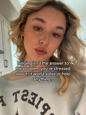 A woman with wavy blonde hair is centered in the frame, looking towards the camera. She's wearing a light gray sweatshirt with visible text, and the background suggests a domestic setting, possibly a kitchen. The lighting is soft, highlighting her facial features.