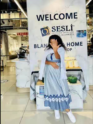 A woman in a blue and white patterned dress and white jacket poses inside a store. She is partially obscuring her face with her hand. Behind her is a large sign that says 'WELCOME SESLI BLANKETS & HOME SHOP.' The setting appears to be a retail store with merchandise displayed on shelves and tables. The overall composition has bright lighting and a clean, inviting atmosphere.