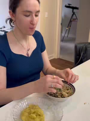 A woman in profile is eating from a bowl. She's wearing a dark blue top, with the food in a bowl placed in front of her. A plate with yellow food is also in view.