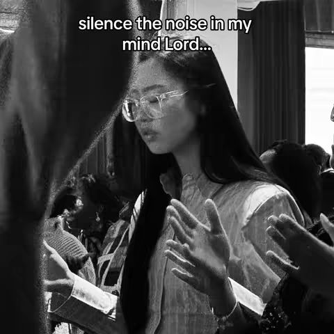 A grayscale image features a young woman with long dark hair and eyeglasses, gazing downward with a contemplative expression. The image has a shallow depth of field, blurring the background and focusing attention on the woman's face and upper body. She's wearing a long-sleeved shirt or blouse, and two other figures are visible in the blurry background, one of whom is partly covering the left of her body. The image has likely been edited to appear monochrome to focus the attention on the main character.