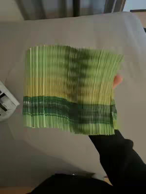 A hand holds a thick stack of folded green and yellow money in front of a neutral background, the arm and hand of the person are visible, with a black sleeve visible.