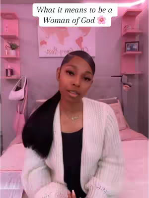 A woman with dark hair pulled back in a ponytail is in a bedroom with pink decorations. She is wearing a white sweater over a black top and looking towards the camera. Above the frame, there is white text, and the background includes a bed and what appears to be a world map.