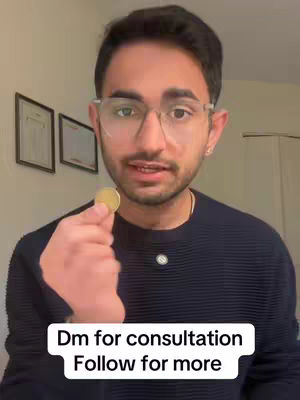A man with glasses holds up a gold coin, looking directly at the camera as if explaining something. Text overlayed on the bottom of the image says 'Dm for consultation' and 'Follow for more.' The background includes framed pictures.