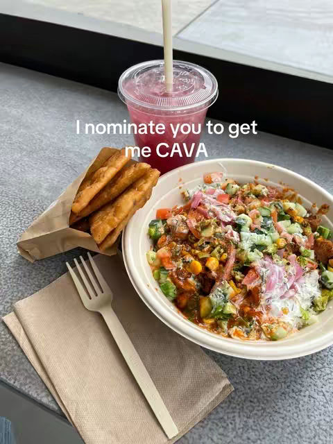 The image shows a close-up of a CAVA bowl, pita chips, and a pink drink. The CAVA bowl is in a white bowl and has a variety of colorful ingredients. A fork rests on a beige napkin beside the bowl, and the pita chips are in a brown paper bag. The pink drink has a clear lid with a straw. In the background, a black surface and a window can be seen.