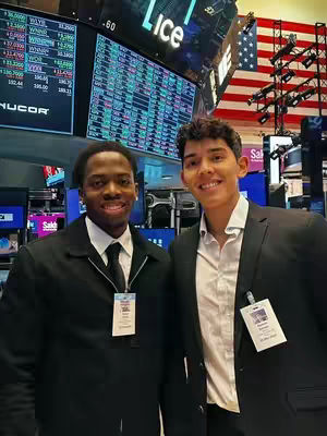 Two young men pose smiling in what appears to be a stock exchange trading floor. One is African American, wearing a dark suit with a black tie, and the other is Latin American, wearing a suit with an open white collared shirt. The background features stock market information displays, the American flag, and other business-related paraphernalia.