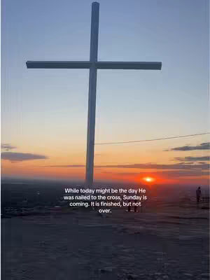 A large white cross silhouetted against a sunset with a person standing in the frame. The overall composition is a landscape with a dramatic sky.