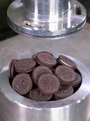 A close-up shot of a pile of Oreo cookies inside a metal bowl. Above, a circular metal fixture is visible, appearing to be part of some industrial process or machine. The arrangement of the cookies and the metallic surroundings create a clean, somewhat sterile, aesthetic, with good lighting highlighting the textures.