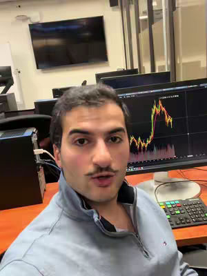A man with a mustache is standing in front of a computer screen. He is wearing a gray zip-up sweater and looking at the camera with an open mouth. The background reveals an office setting with multiple computer screens, a desk, and a TV screen.