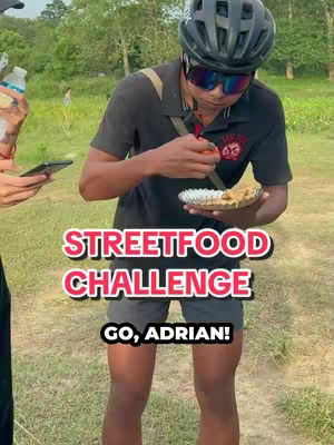 A man in a black shirt, bike helmet, and sunglasses stands on a grassy area, holding a small plate of food. The background shows trees and a clear sky and some people. Overlay text in the image reads "STREETFOOD CHALLENGE" and "GO, ADRIAN!"