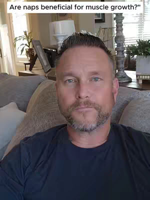 A man with short, dark hair and a trimmed beard sits on a couch. The background is a living room setting with a lamp.