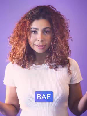 A woman with long, curly auburn hair is centered against a light purple background. She is wearing a white t-shirt and has a small blue rectangle overlaid on her chest with the word 'BAE' in white. She has a friendly expression.