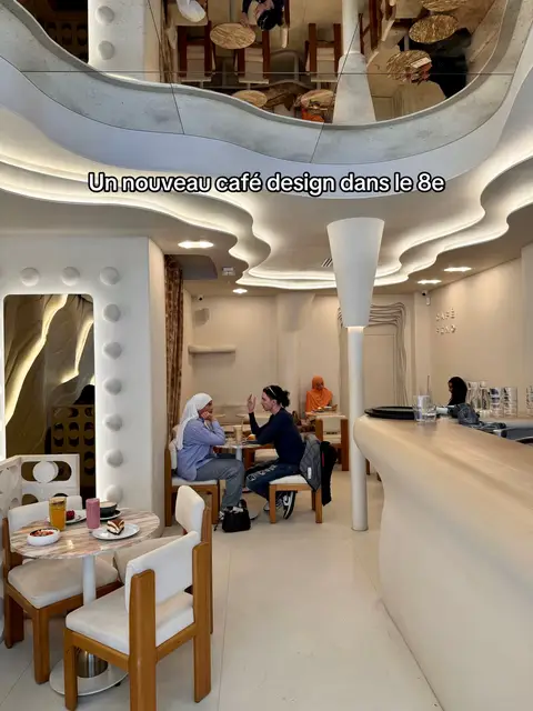 The cover image shows the interior of a modern-designed cafe. The space is primarily white with curved architectural elements and a reflective ceiling. Two people are seated at a table, engaged in a conversation with animated gestures and they seem to be smiling. Another person is in the background, and the layout includes tables, chairs, and a curved bar, suggesting a bustling cafe atmosphere.