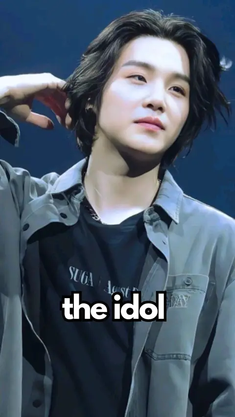 A close-up shot of a man with dark hair, gazing slightly upwards and to the side. He has pale skin and is wearing a gray, open shirt over a black t-shirt. The t-shirt features the word "SUGA" and some other text. The background is a blurry blue. The text overlayed on the image reads "the idol".