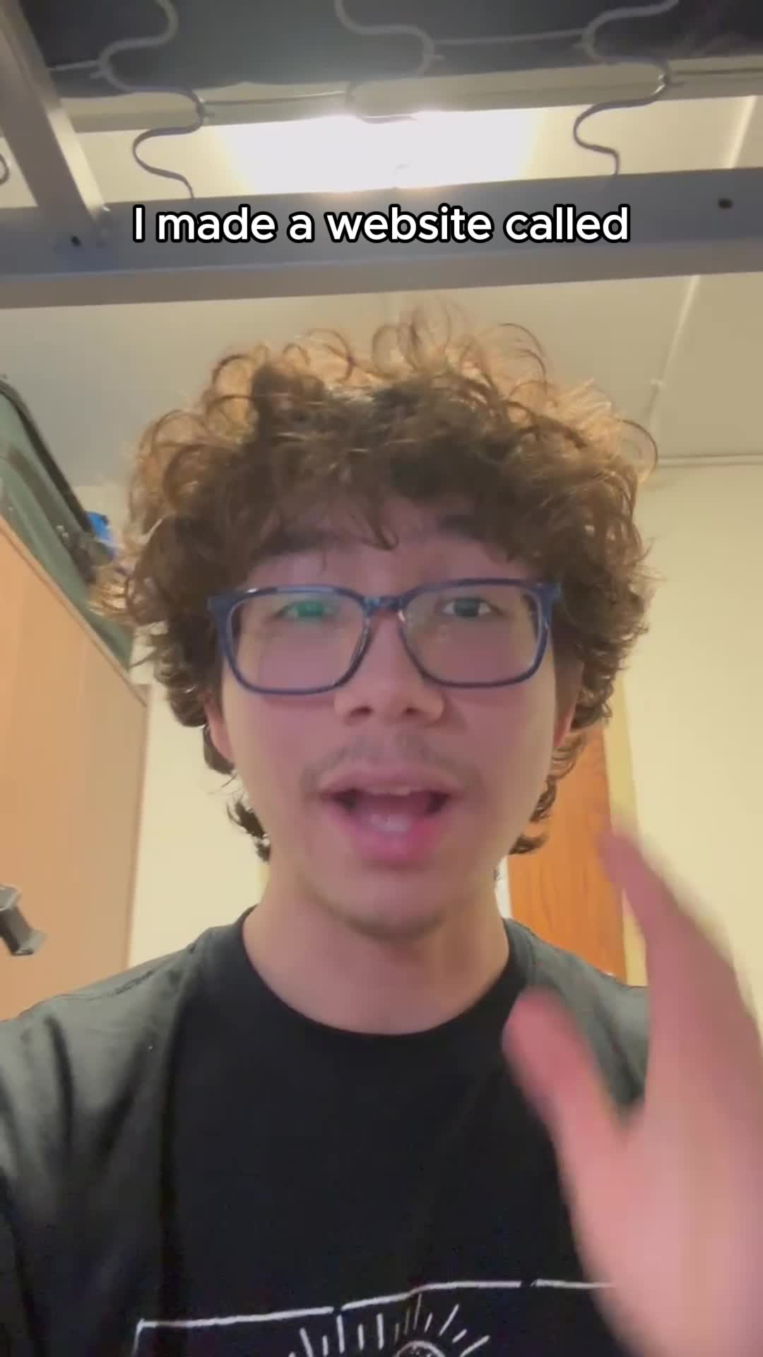 A young man with curly brown hair, wearing glasses and a black t-shirt, is speaking to the camera. The background shows a plain wall and part of a bed frame. The top of the image has a semi-transparent white box with the text overlayed