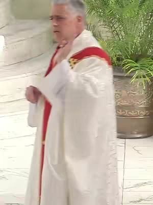 The image captures a man in a white religious garment with red accents. He is seen from the waist up, with a slight tilt of his head to the side. The background shows part of a stone wall and a touch of greenery in a decorative pot, suggesting an indoor architectural setting.