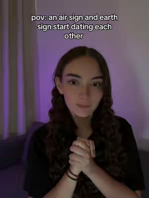 A woman with long brown hair and a black shirt is centered in the frame. She clasps her hands together in front of her chest. Text overlayed on the image reads 'pov: an air sign and earth sign start dating each other'. The background is a blurred purple color, and the overall composition is focused on the woman's upper body and face.
