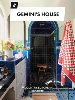 The image showcases a luxurious bathroom. The shower is tiled in dark blue with a wooden stool inside and a glass door. Adjacent to the shower, a white cabinet with drawers holds various items. A red and white checkered towel hangs, providing a pop of color against the blue. The text overlay indicates the content is related to a Gemini's house decor, described as country European and rustic style. The composition is framed by vibrant blues, greens and whites.