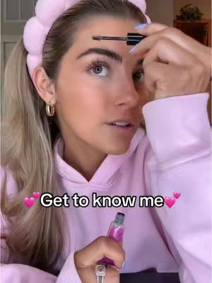A young woman is holding a makeup product in one hand and a mascara wand in the other, in the process of applying makeup. The text overlay shows 'Get to know me'. She wears a pink hoodie, a headband, and hoop earrings. The background is blurred and appears to be indoors.