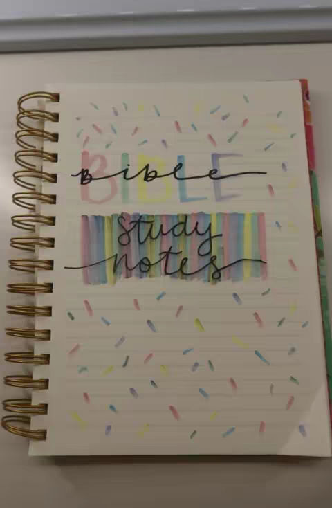A notebook with a gold spiral binding is open on a white surface. The pages are lined and have a white background with colorful confetti-like speckles. The words "bible" and "study notes" are handwritten in black marker over a colorful, striped background, providing a visual accent.