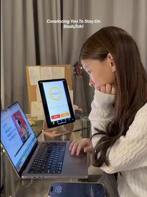 A young woman sits at a desk in a dimly lit room, focused on a laptop and a tablet. The laptop displays a website, while the tablet shows a timer. A phone rests on the desk. The image is composed with the woman in profile, her hand supporting her chin.