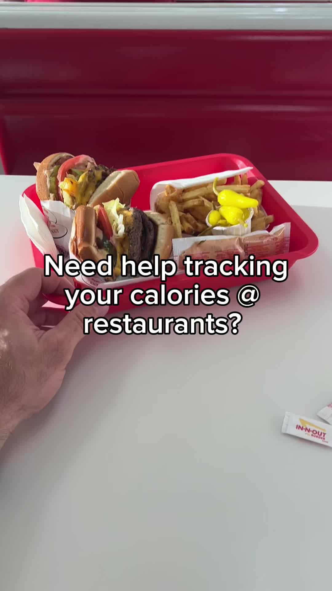 A person's hand holding a red In-N-Out tray with a burger, fries, and condiments. The background has a red booth.