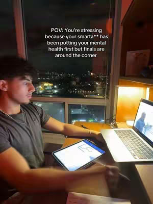 A young man is seated at a desk near a window, looking towards a laptop display. The interior has a warm tone due to the lighting. The lighting suggests a night-time setting.