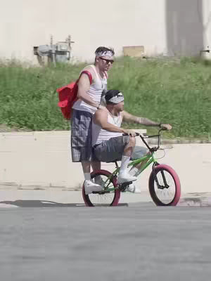 Two men are outside on a sunny day. One is sitting on a green and pink BMX bike, while the other man is standing next to him, appearing to be helping him. The setting is outdoors, with a neutral-toned wall on the right side and green foliage in the background.