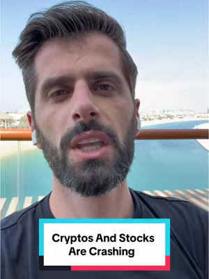 The video cover features a man in a close-up shot. He is wearing a black t-shirt and white earbuds. The background shows a bright, sunny outdoor scene with water and buildings. A text overlay at the bottom says, 'Cryptos And Stocks Are Crashing,' set against a teal and pink rectangular background.